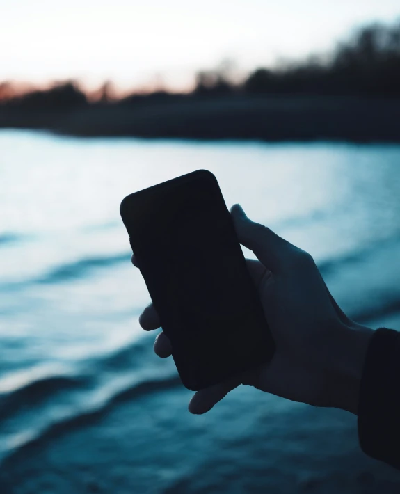 person holding up a phone near water with sunset in the background