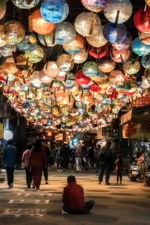 people sitting in the middle of a street with multicolored paper lanterns above them