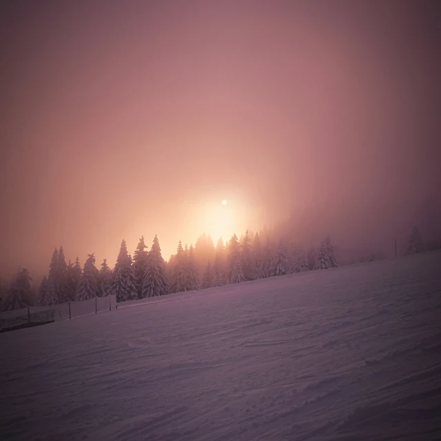 a snowy mountain has trees and the sun shining through the clouds