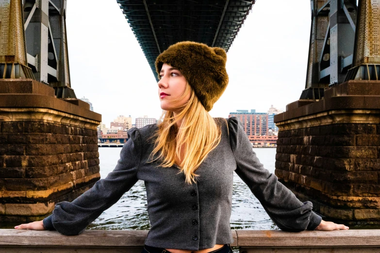 a blonde haired woman wearing a furry hat stands by a bridge