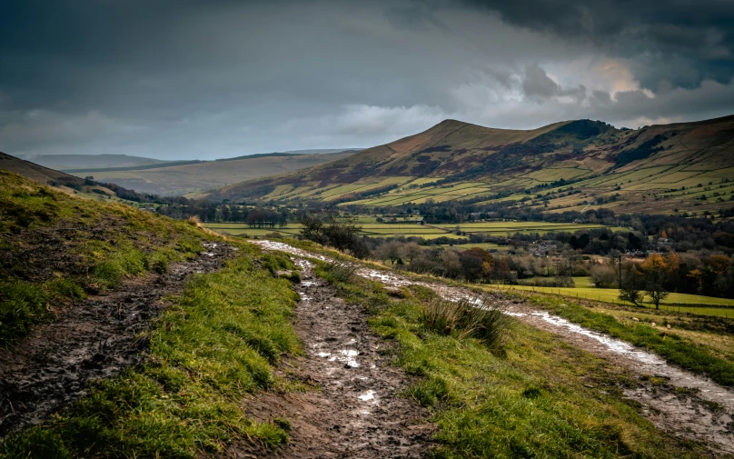 a muddy dirt path splits into a grassy field with rolling hills in the distance