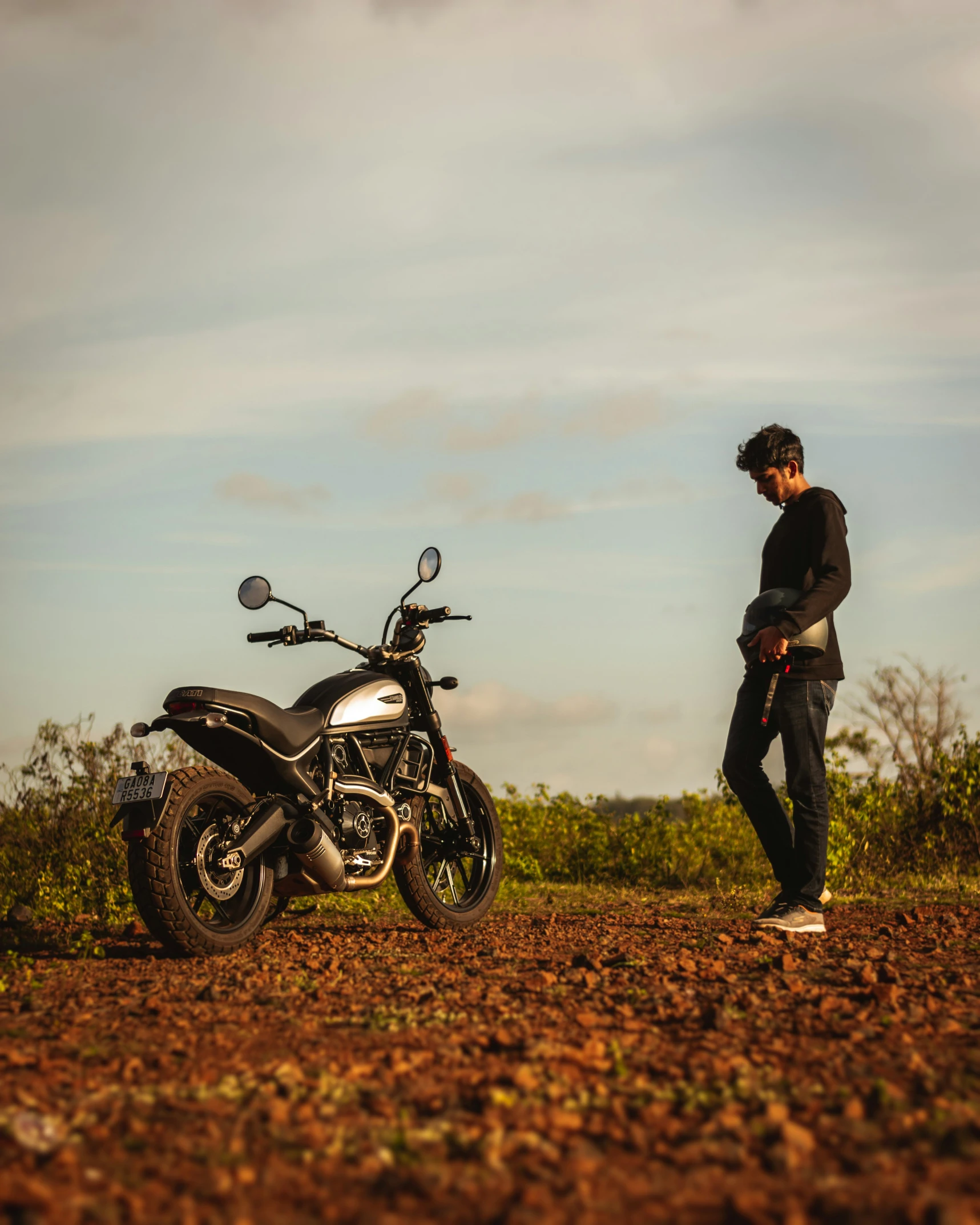 a young man stands by a black motorcycle in a field