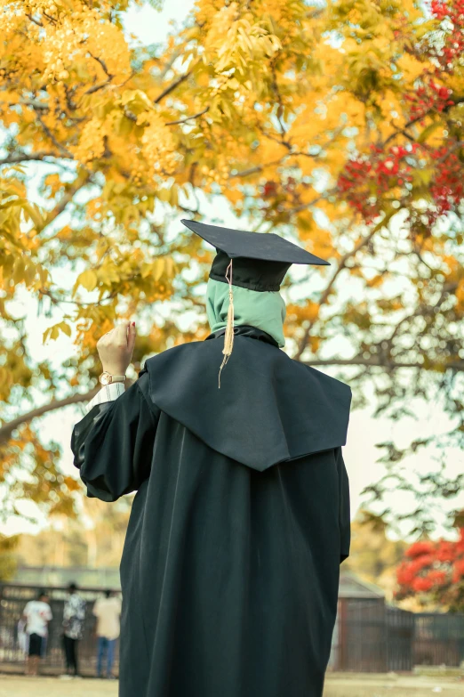 a person in black graduation gown and mortar cap stands in front of a tree with leaves and yellow foliage