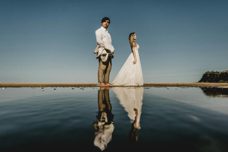 a bride and groom standing on the water reflecting the clear sky