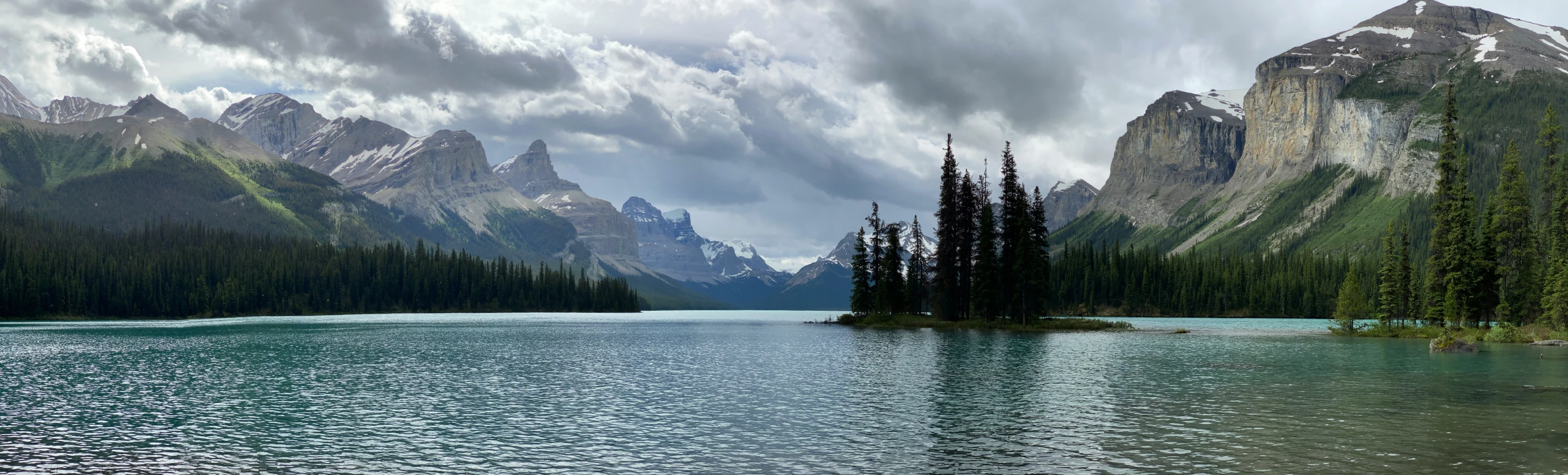 clouds hover above some mountains as trees stand in the water