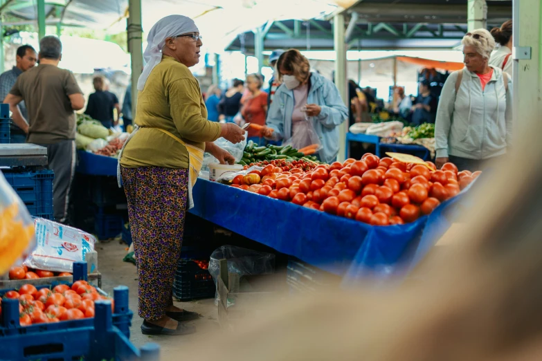 a woman standing in a market with a big blue container filled with tomatoes