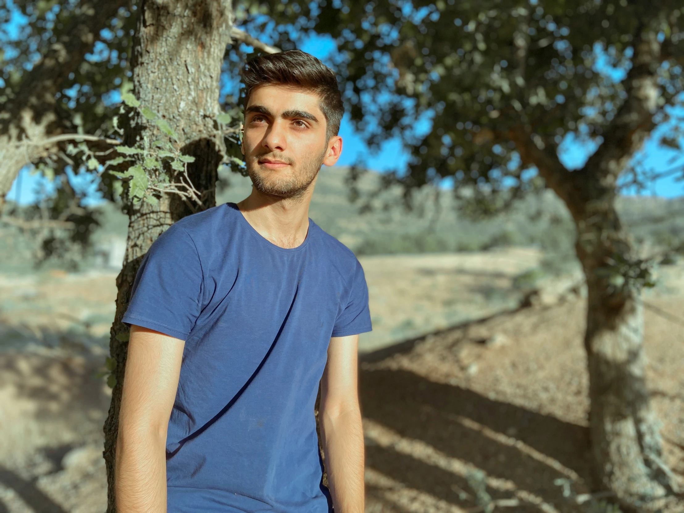 a man with dark hair in blue shirt near trees