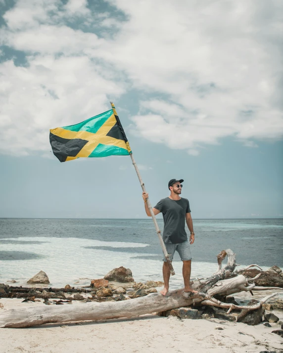 man holding jamaica flag while standing on driftwood by beach