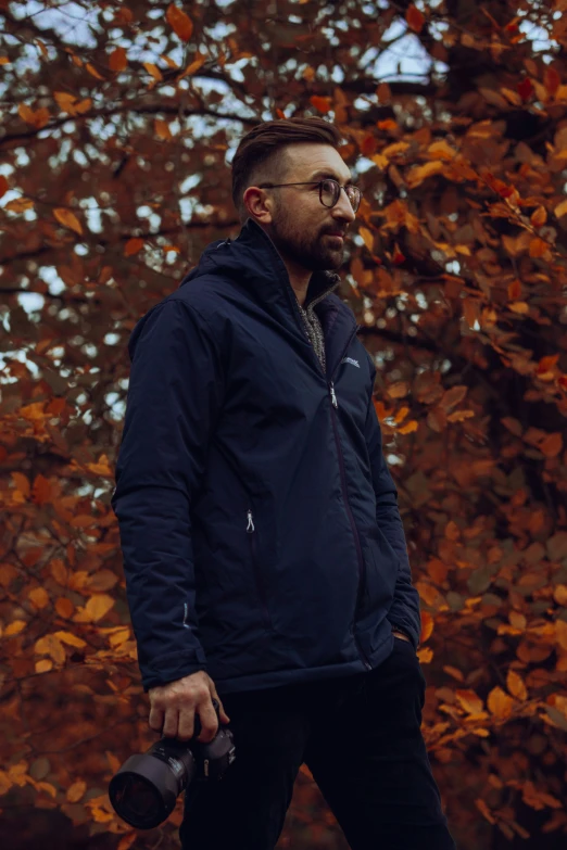 a man with a beard wearing glasses, blue jacket and holding two camera equipment