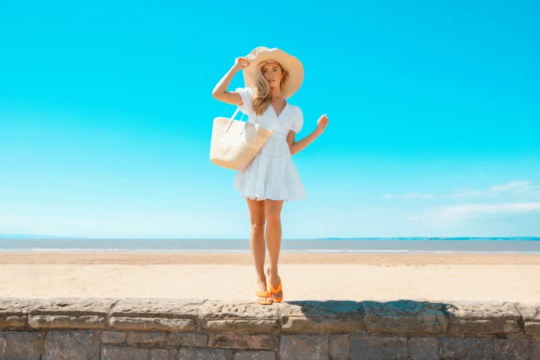 a woman in a white dress and hat holding a tote bag is standing on the wall next to the beach