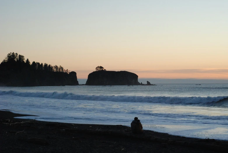 someone sitting on the shore watching a large rock in the ocean