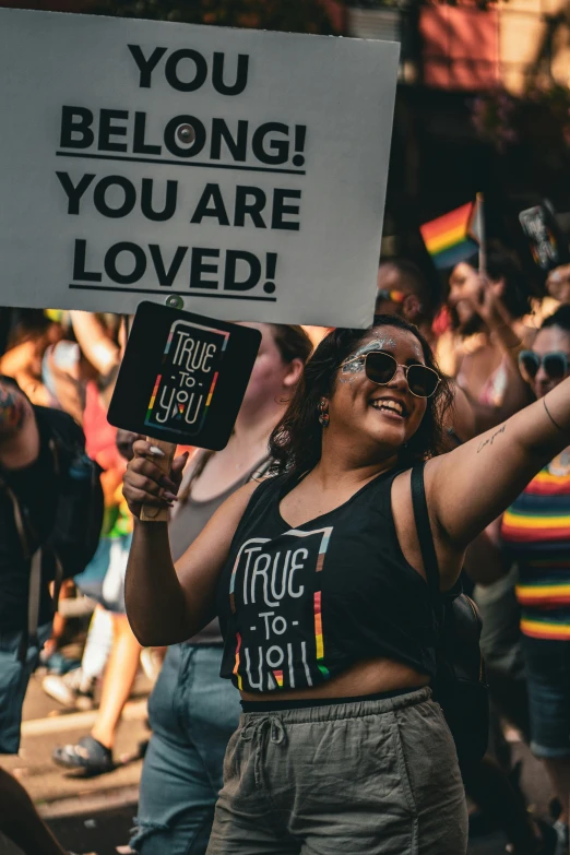 people gathered at a gay pride event while one girl holds a sign reading you belong, you are loved