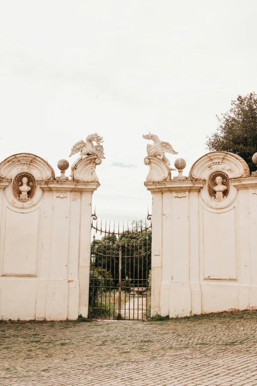 an open gate with statues above it in front of a stone driveway