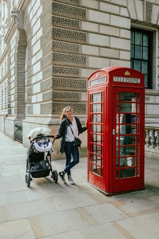 a woman with a baby stroller hing a baby carriage past a red telephone booth