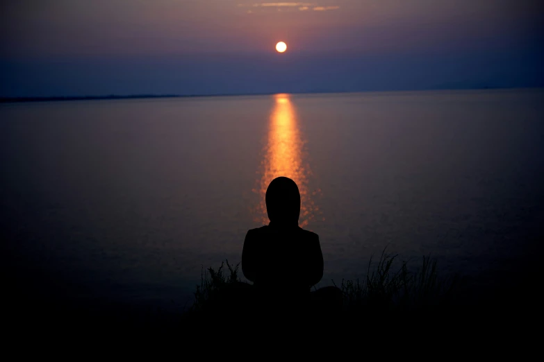 a person is sitting down near the ocean looking at a setting sun