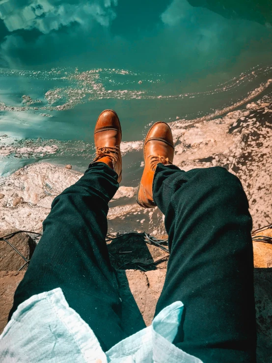 person with brown shoes sitting on rock looking over body of water