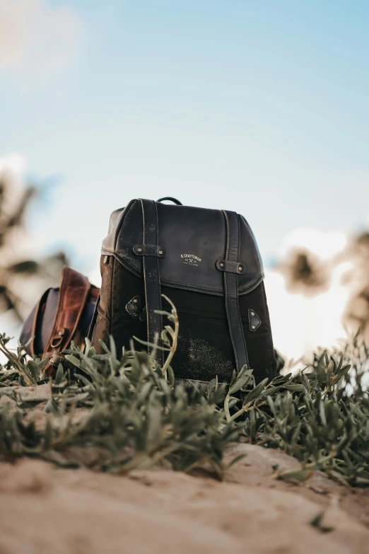 two brown leather backpacks sitting on sand and grass