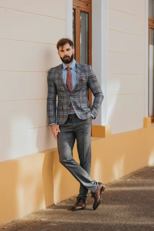 a man in a plaid jacket and tie leaning on a wall