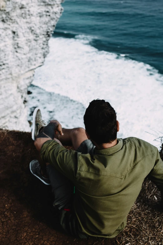 a man is sitting at the edge of a cliff overlooking a body of water