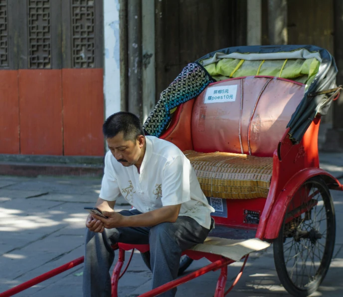 man sitting on red cart using cellphone near brick wall