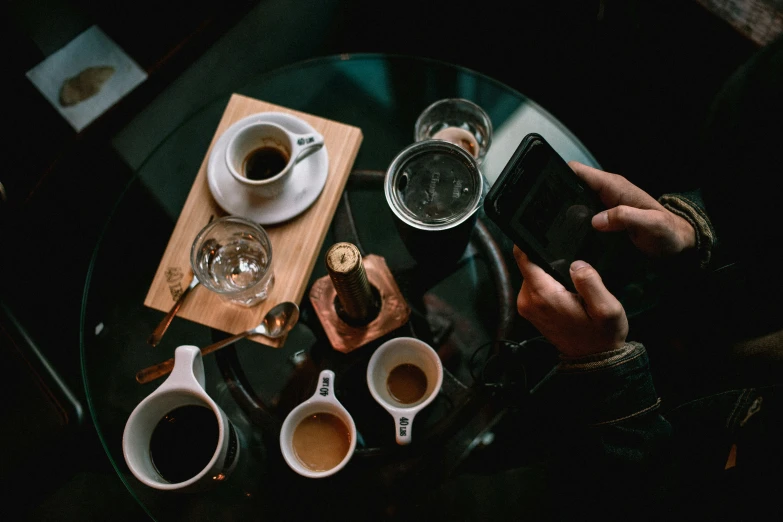two cups of coffee sit on a glass table next to a phone, with some wine glasses, an espresso can and other cups on the table