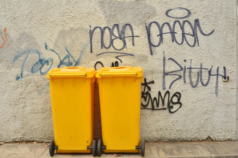 two yellow garbage cans next to a wall with graffiti