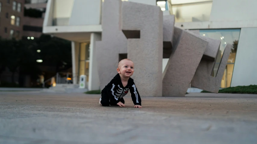 a smiling toddler sits on the cement floor near some modern buildings