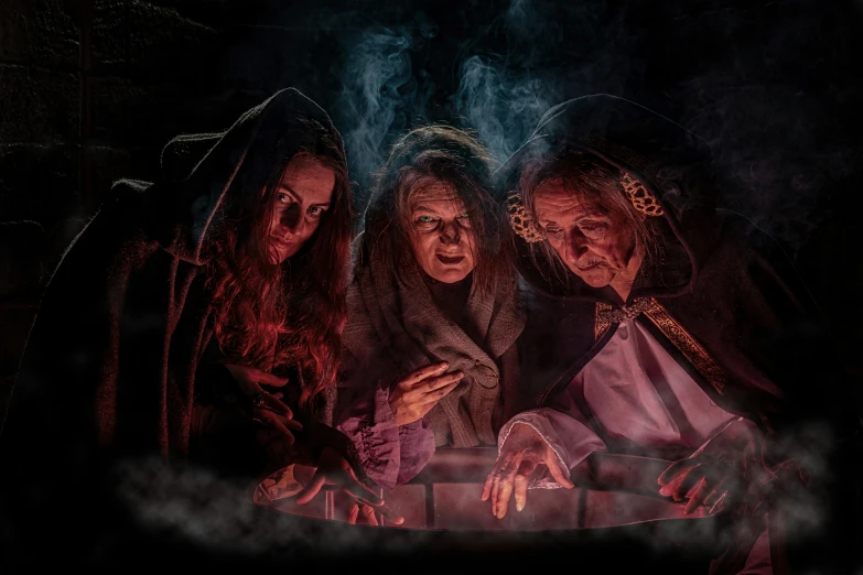 three witches gathering over a table in the dark