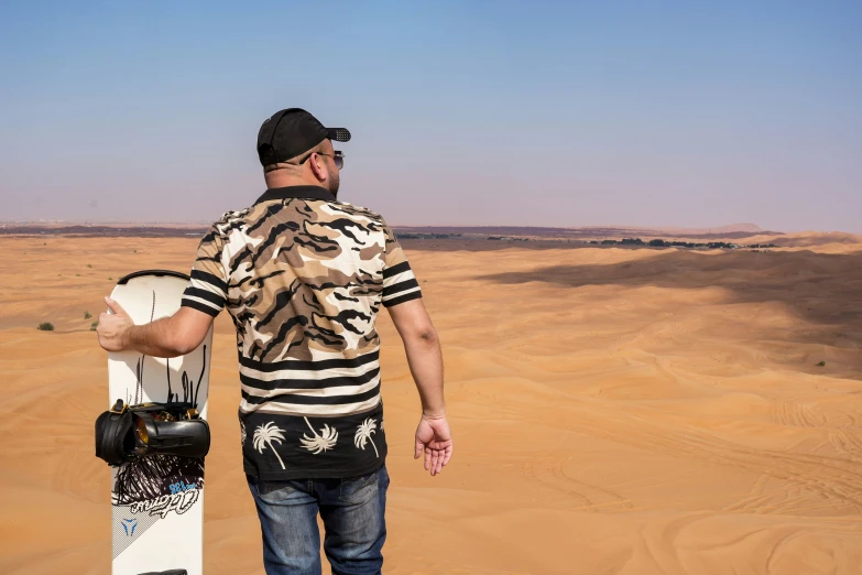 man standing on desert, wearing an animal print shirt with his hand on a skateboard