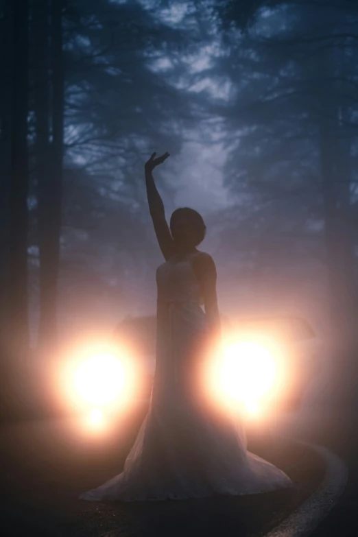 a woman stands in the middle of a path holding her arm up in the air with two headlights in front of her