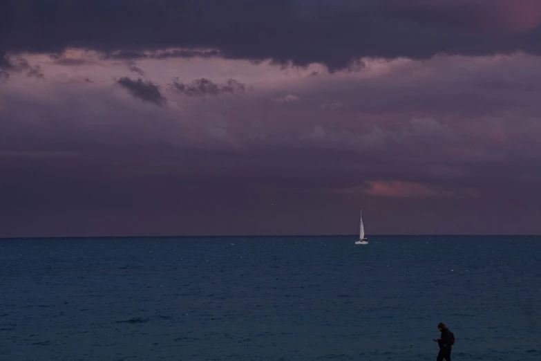 the person is standing on the beach at dusk with a sailboat in the distance