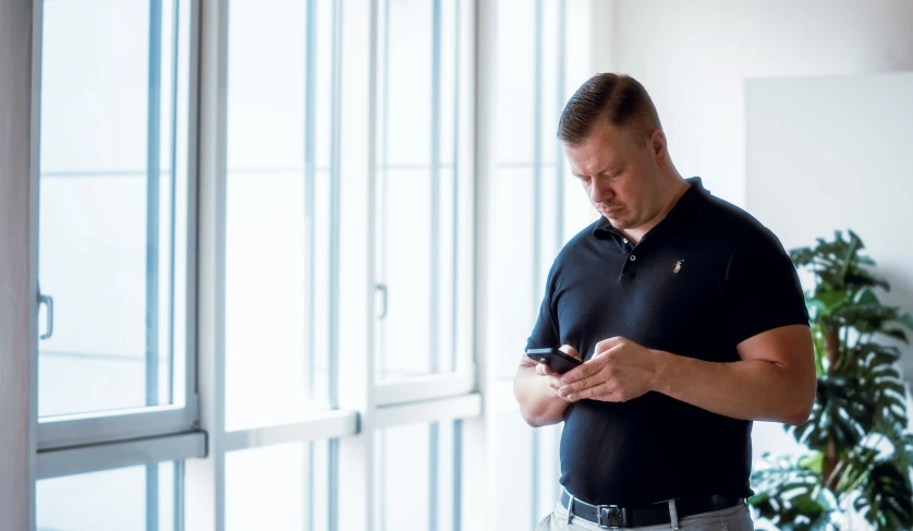 the man is standing near a window looking at his phone