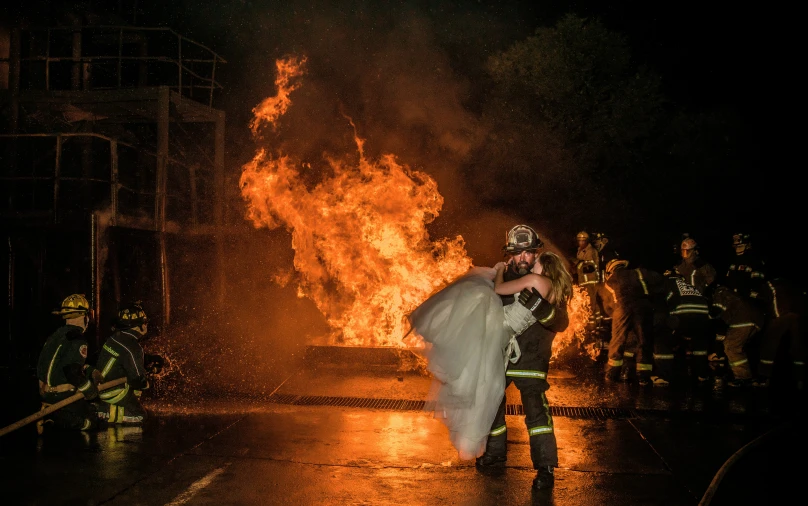 a man standing in the rain holding a wedding dress next to a fire