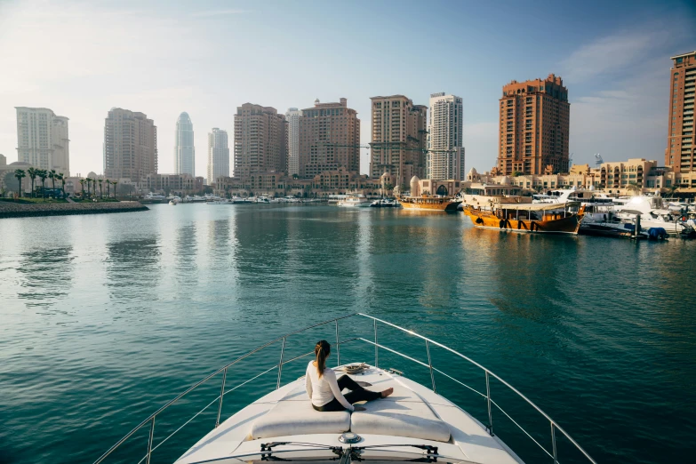 a woman sits on the bow of a boat with boats in a harbor near skyscrs