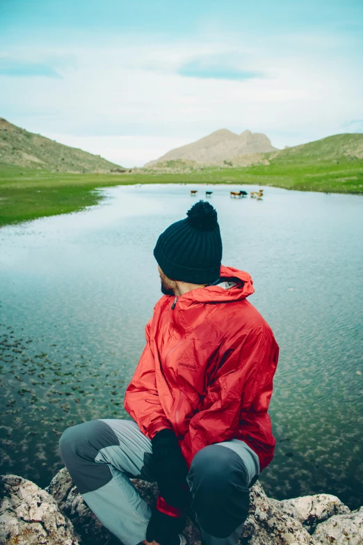 a person sits near a small pond wearing a hat