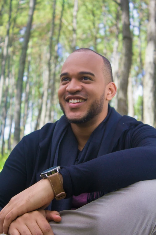 a smiling man in a sweater sits among the trees