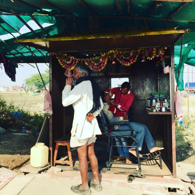 the man wearing his sandals is taking a picture while another mans sitting outside of a hut