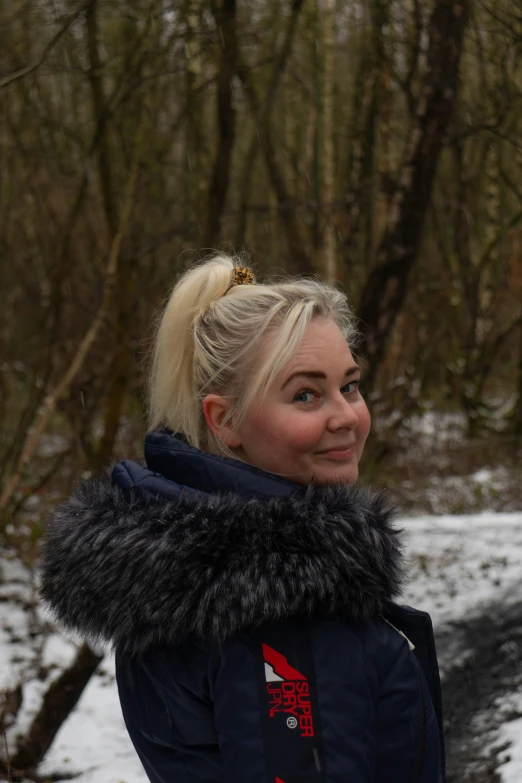a blonde woman wearing a coat and a scarf smiles to the camera
