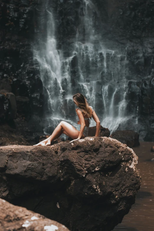 a woman in swimsuit sitting on rock next to waterfall