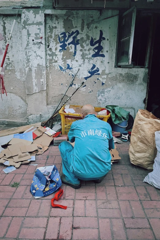 the back of a man squatting on a cobblestone sidewalk, with an assortment of boxes and other items piled on the ground