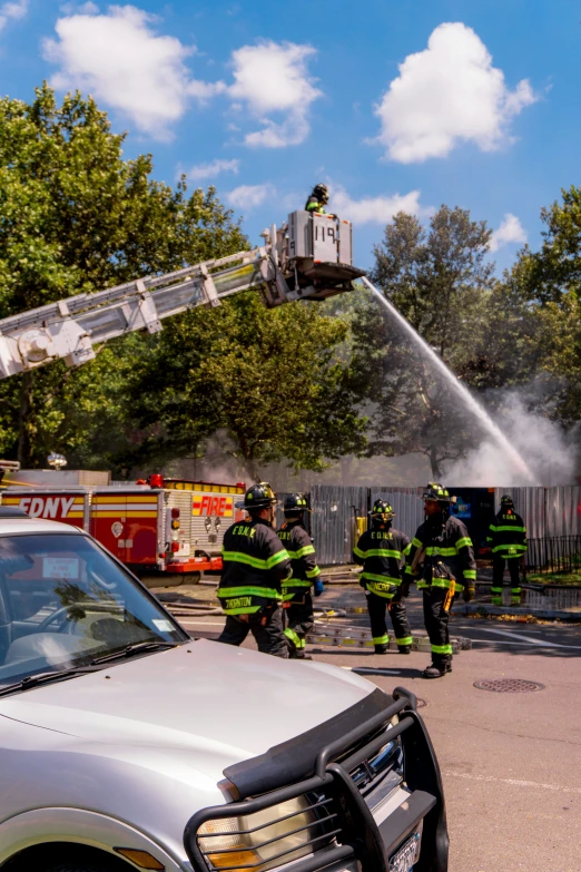 two firemen watching a car go through a fire with a hose on it