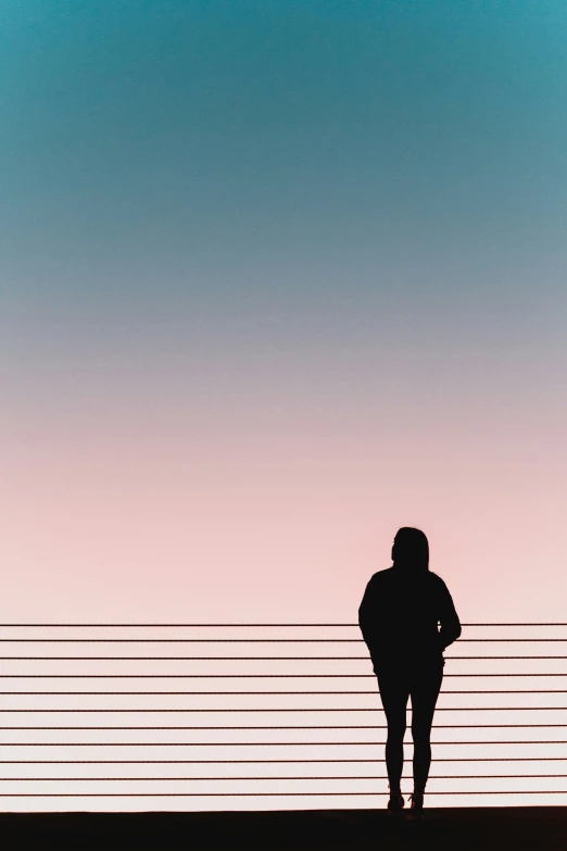 the silhouette of a person standing by a fence at sunset