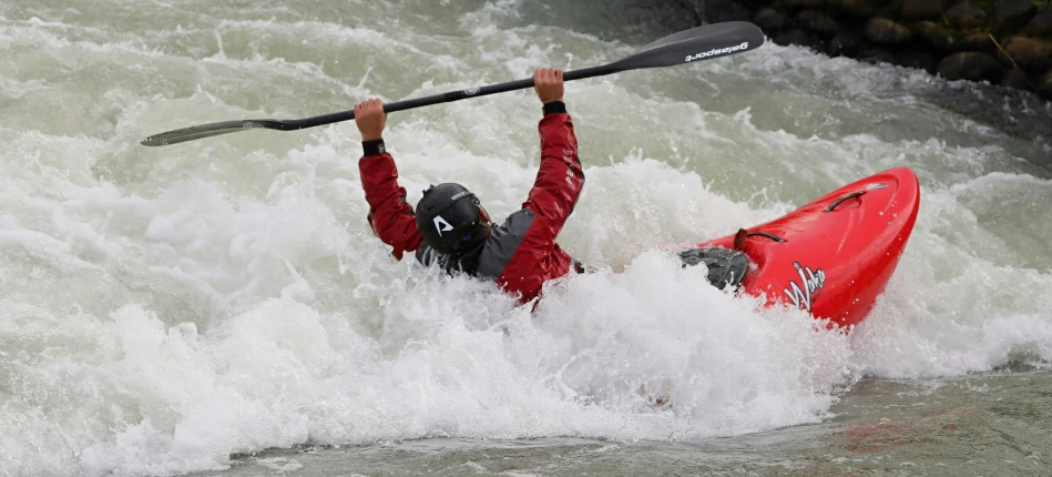 a man wearing a red wetsuit paddles through a wave on a kayak