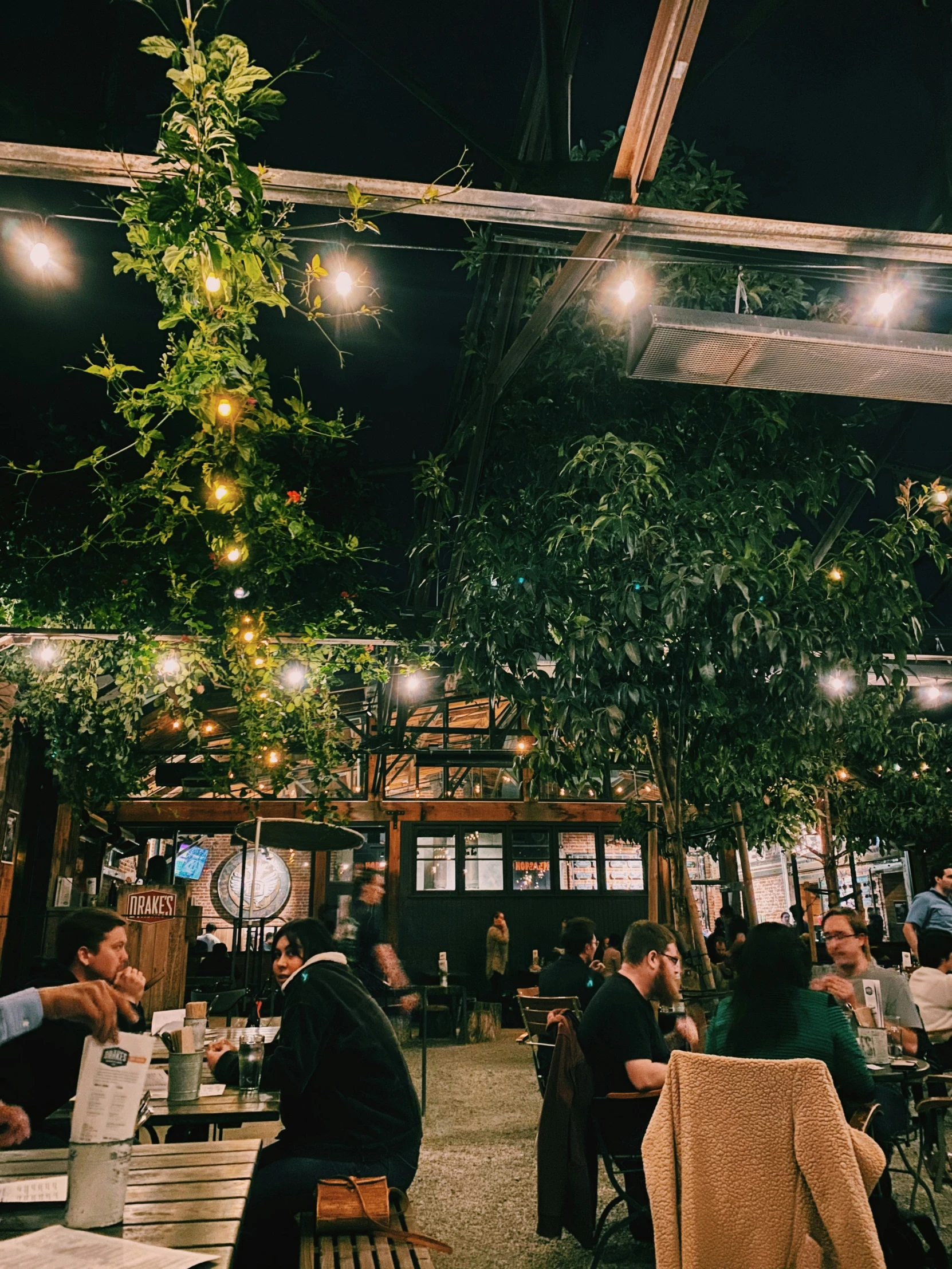 the inside of a restaurant with long tables, hanging lights and lots of people sitting around it