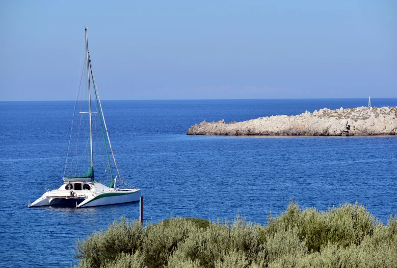 a sail boat docked on the ocean in front of a beach with trees and rocks