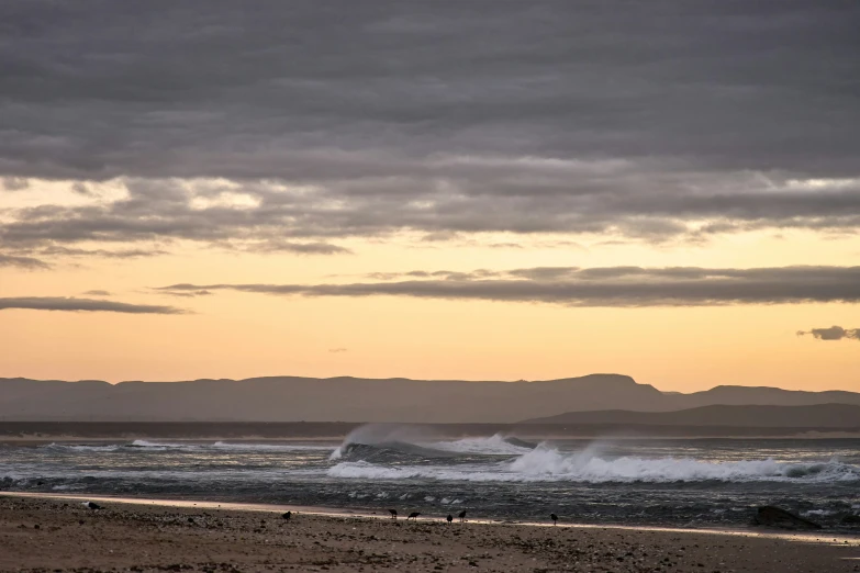 this is a beach at sunset, with waves crashing on the shoreline and mountains in the background