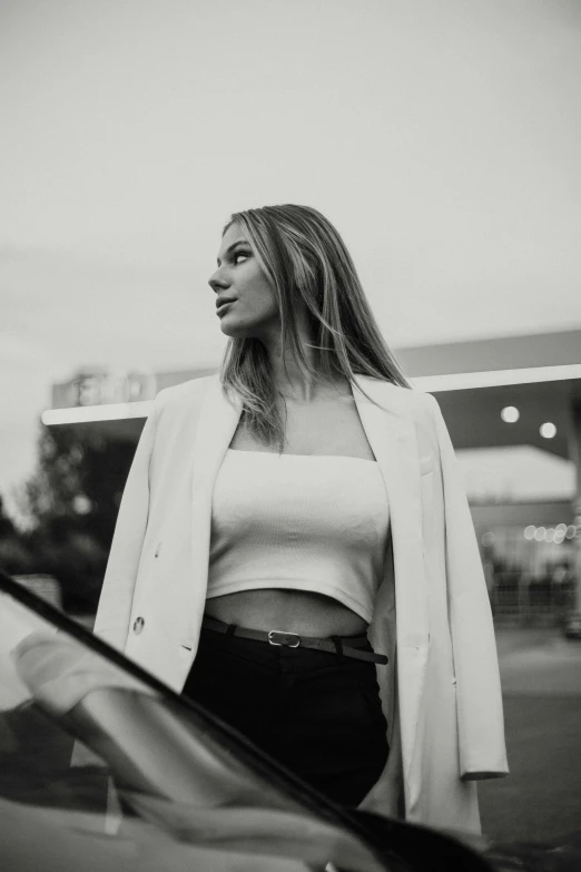 a woman in a crop top and blazer leaning on the hood of a car