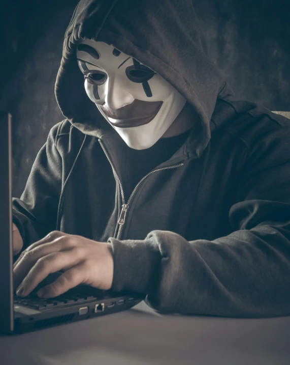 the person is wearing a creepy mask on their face, and typing on a laptop