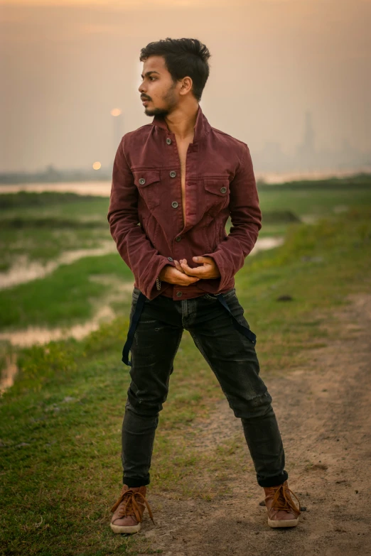 a young man wearing a maroon jacket holding a brown leather object in one hand and his hand over his chest, standing on a dirt path