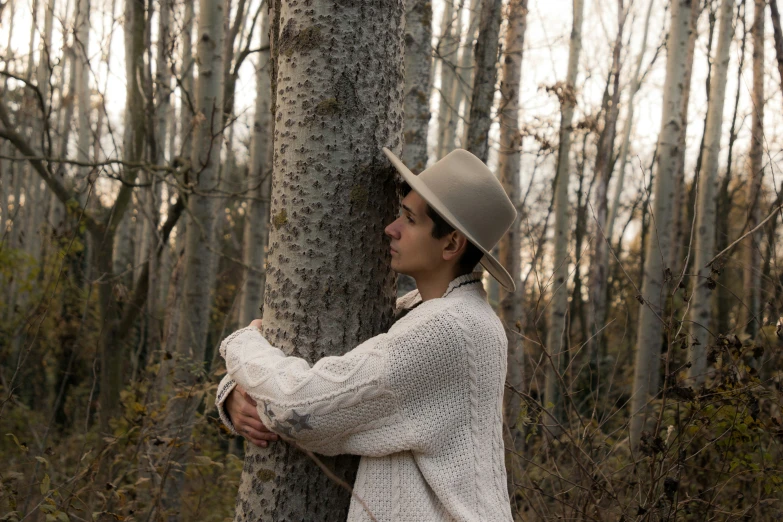 a boy leans against a tree and wears a fedora